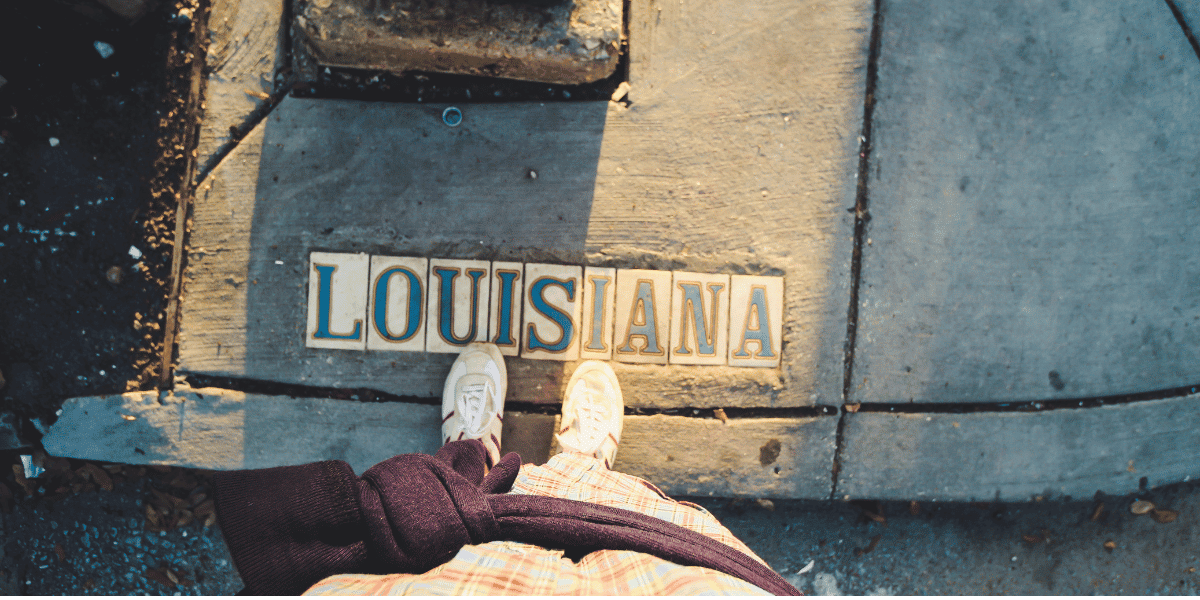 Person standing on a street corner looking down at traditional Louisiana street marker tiles that spell out Louisiana