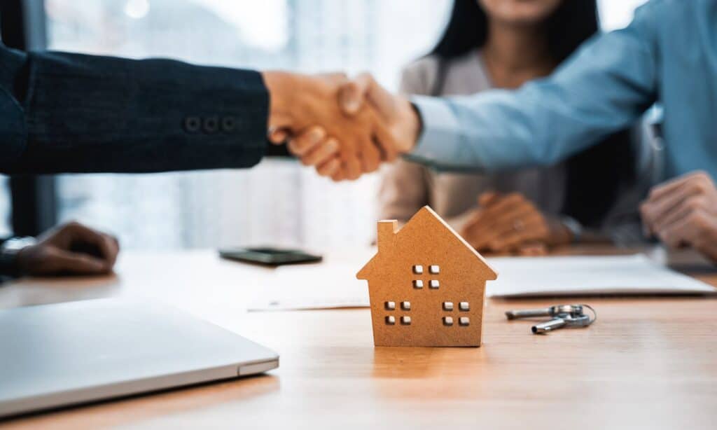 Two people shaking hands over a table with a house model and keys, while a couple of keys post-sale