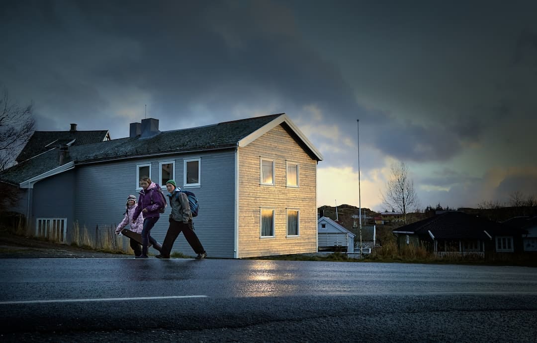 A female real estate agent professionally dressed gestures while talking to a young couple inside a bright, modern living room with large windows.
