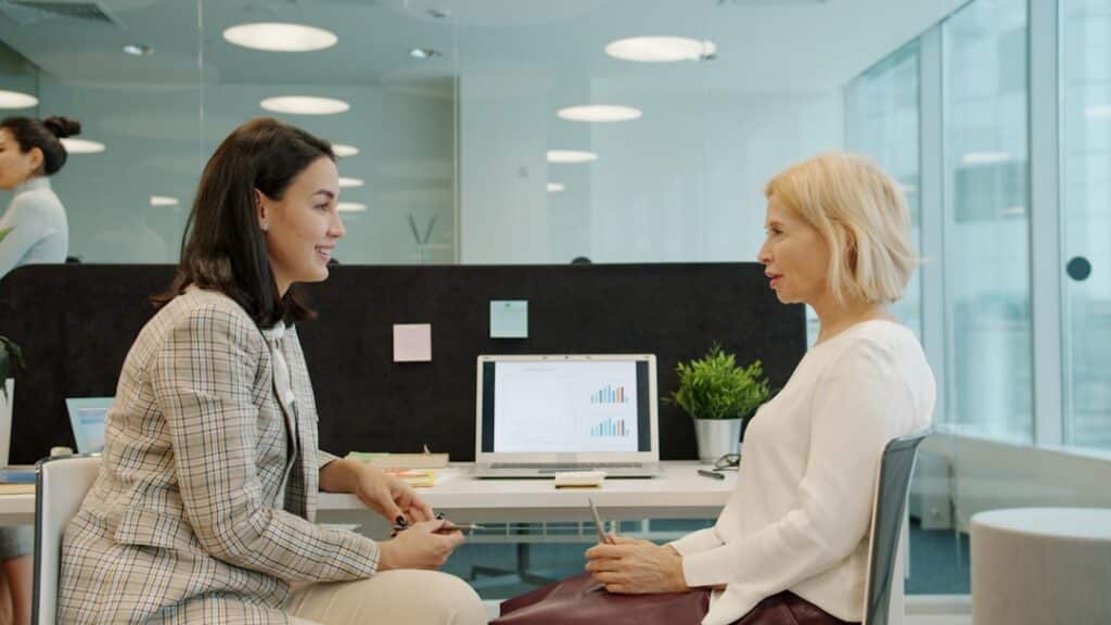 Man and a woman in business casual attire sit opposite each other at a minimalist wooden desk, engaged in a serious and pr...