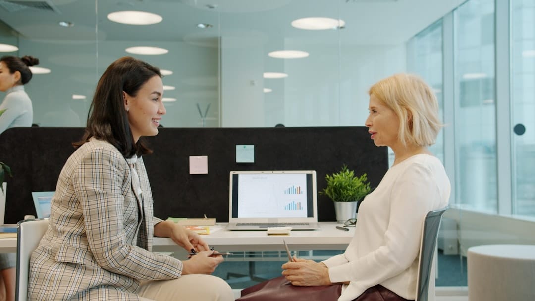 Man and a woman in business casual attire sit opposite each other at a minimalist wooden desk, engaged in a serious and pr...