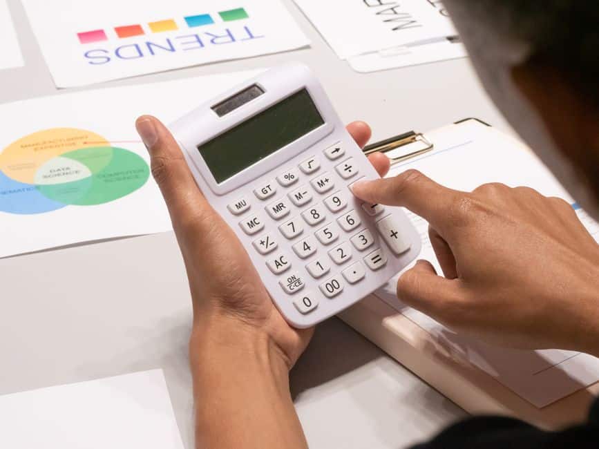 Business professional sits at a bright, modern desk, using a calculator next to a laptop and notepad, carefully planning a...