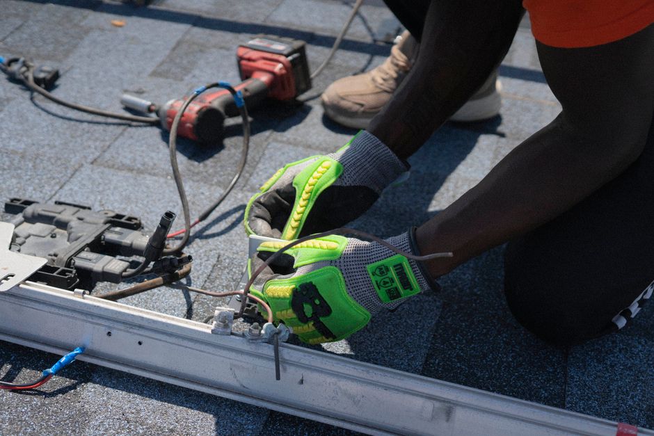 A professional roofer wearing safety gear carefully installs new architectural shingles on a residential roof on a bright, sunny day, representing quality craftsmanship.